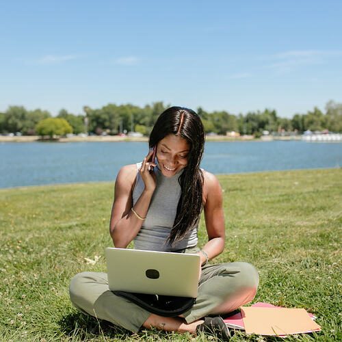 Photo: Pexels, RDNE Stock project Smiling woman sits on grass by a lake, using a laptop and talking on the phone, enjoying the outdoors.